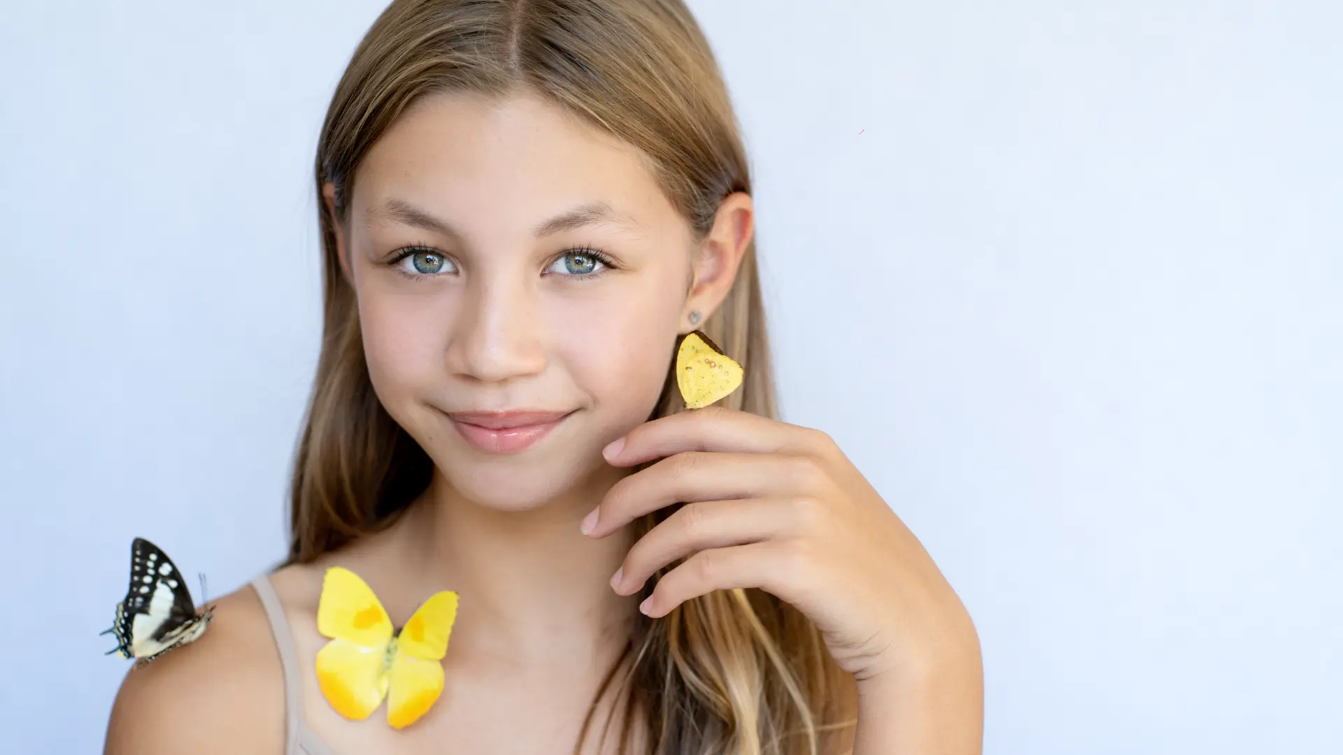 Girl with light-brown hair and yellow butterflies resting on her hand and shoulder, against a pale backdrop